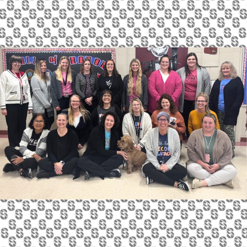 A group of school staff pose together in a hallway, some seated and some standing, with a festive bulletin board behind them.”
