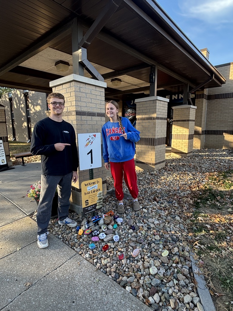 “Two students stand beside a ‘Welcome to our Kindness Rock Garden’ sign with decorated kindness rocks displayed at its base.”