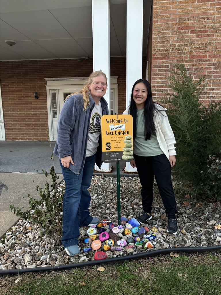 “Two adults stand smiling beside a Kindness Rock Garden sign with colorful painted rocks gathered at the base.”