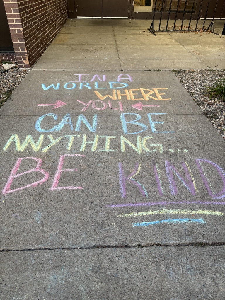 Sidewalk chalk message reading ‘In a world where you can be anything… be kind’ in bright pastel colors outside a school entrance.”