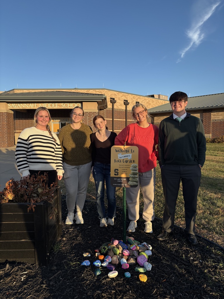 “Five students stand behind a Kindness Rock Garden sign outside Savannah Middle School with painted kindness rocks in front.”