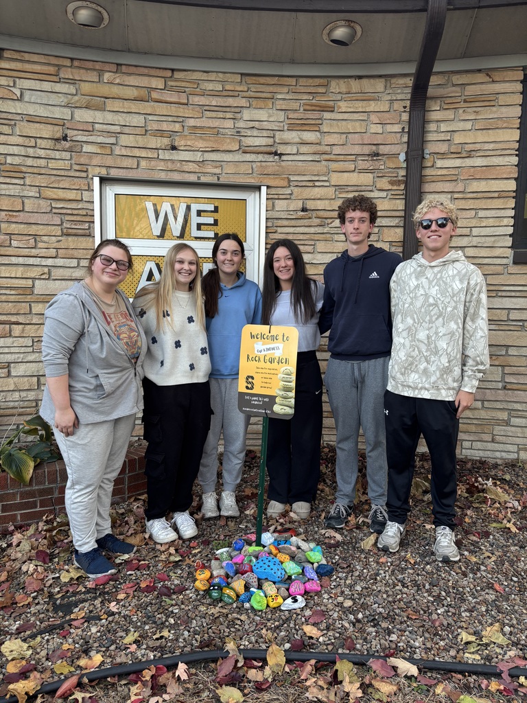“Six students stand around a Kindness Rock Garden sign outside a school, with brightly painted rocks arranged on the ground.”