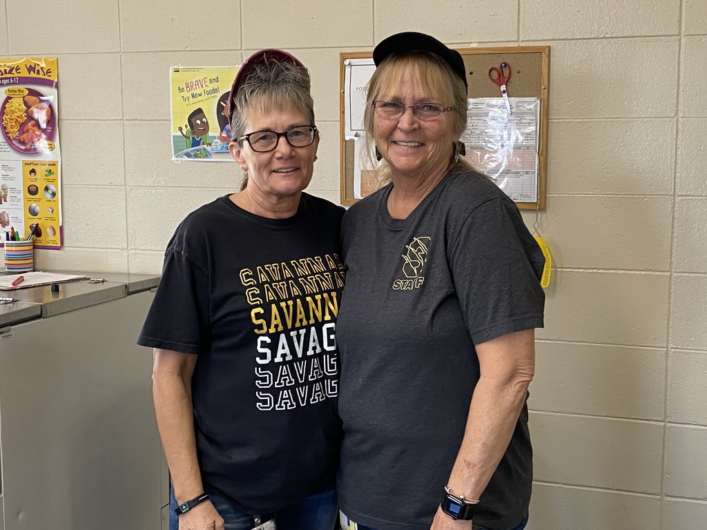 Two kitchen staff members stand together in front of a beige wall and food service area, smiling at the camera.