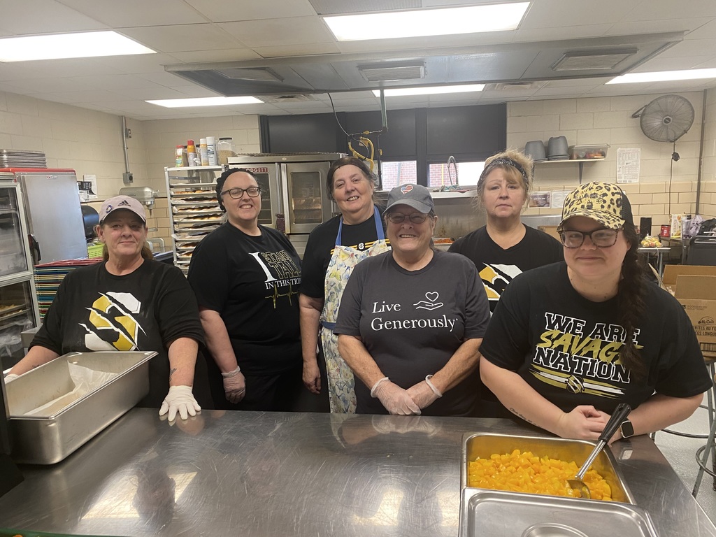 Six kitchen staff members pose together in a school kitchen, standing behind trays of prepared fruit.