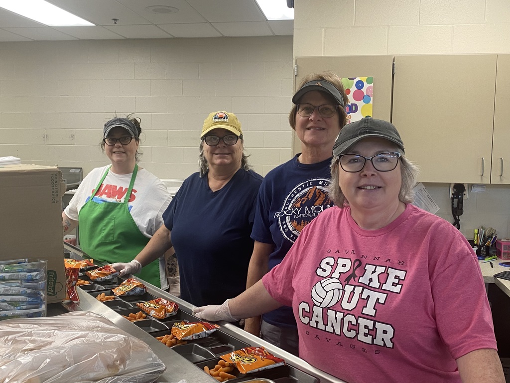 Four food service staff members stand behind a lunch line preparing trays with chips and carrots.