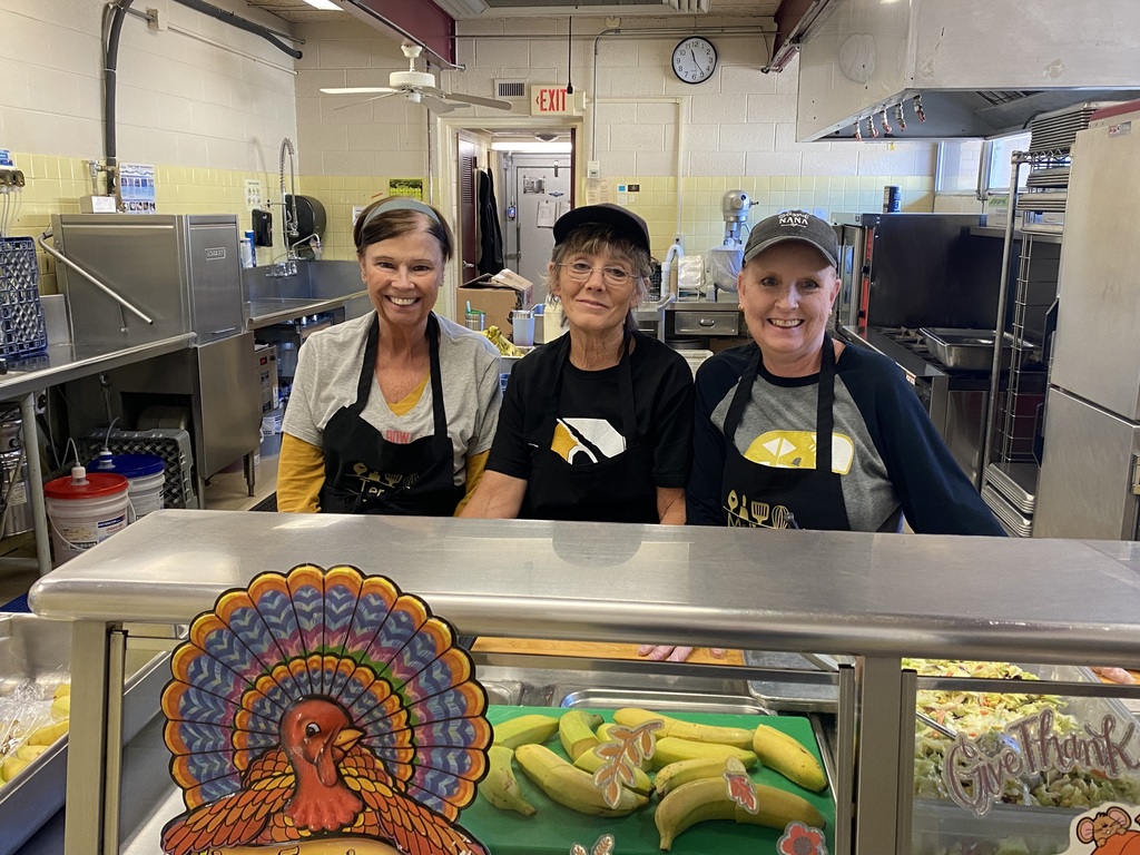 Three food service staff members smile from behind the serving line decorated with Thanksgiving artwork and trays of bananas and salad.