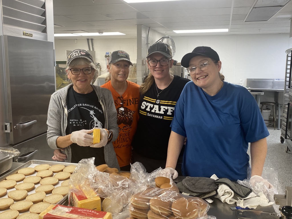 Four food service workers stand behind a counter filled with buns and cheese slices, smiling for the photo.