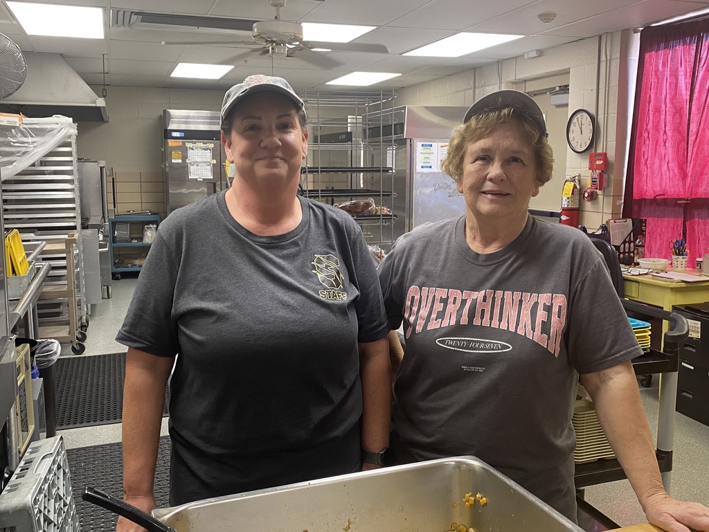 Two kitchen staff members stand side-by-side in a school kitchen near a food pan.