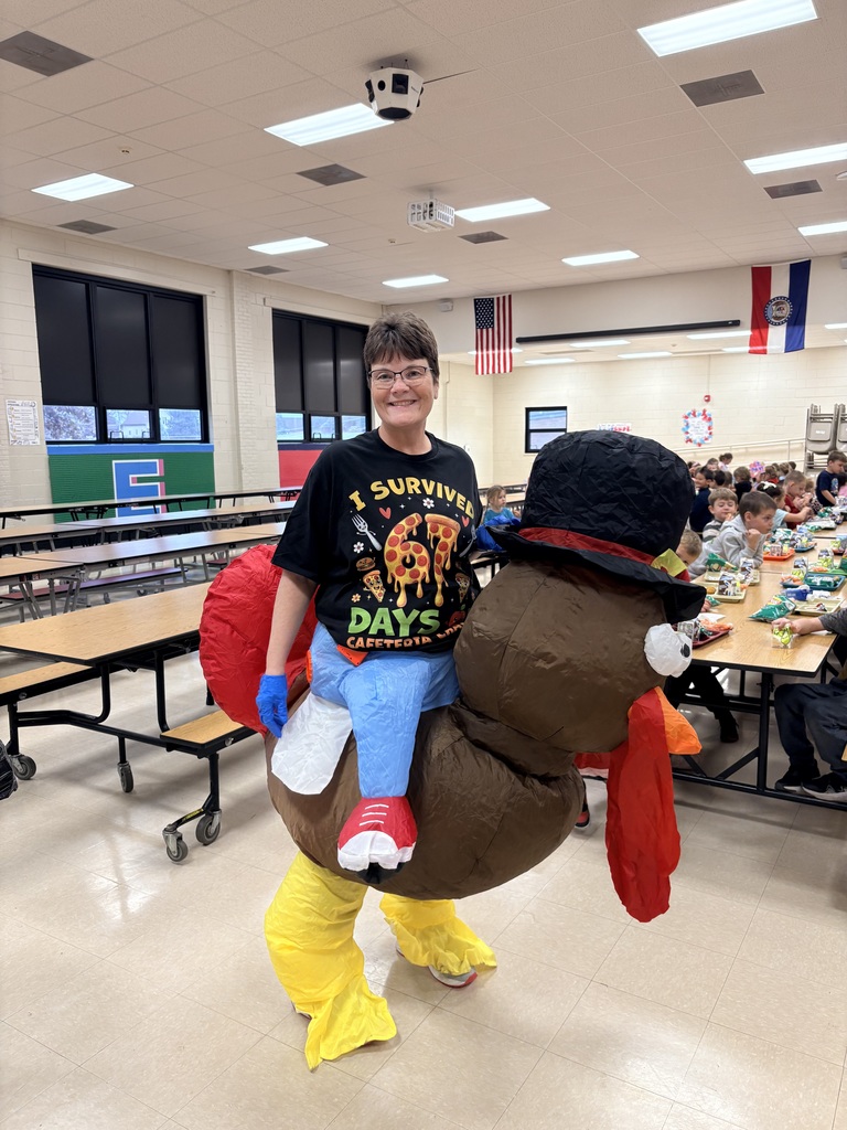 Lunch duty staff member wearing a ‘67 Days of Cafeteria Chaos’ shirt and an inflatable turkey costume in the school cafeteria with students eating in the background.