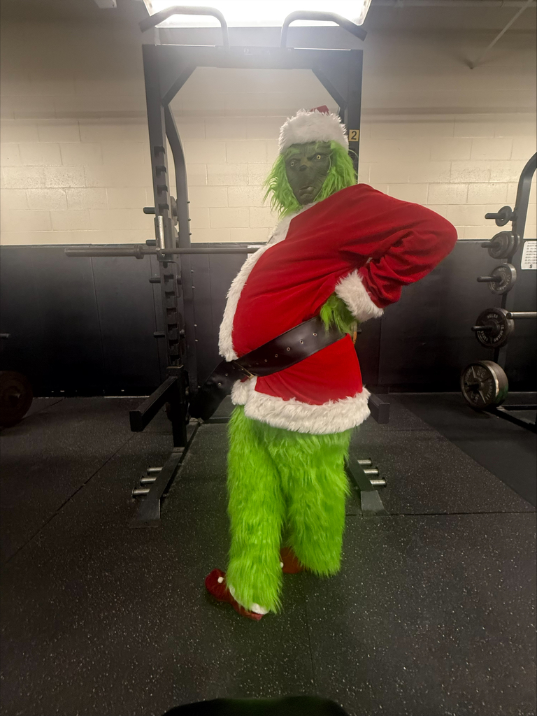 The Grinch in a Santa outfit poses playfully in a school weight room, standing in front of a squat rack.