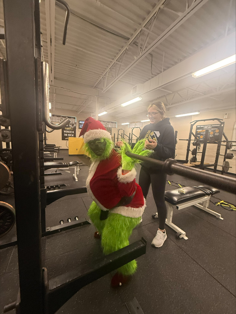 The Grinch leans toward a barbell in the weight room while a smiling student stands behind him.