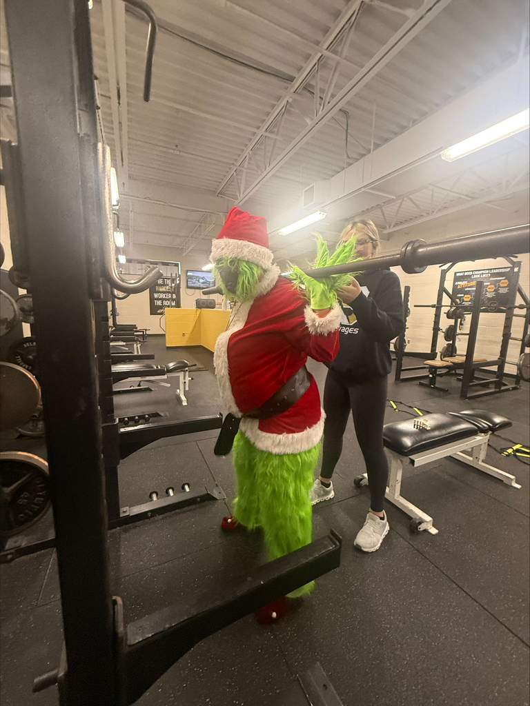 The Grinch stands at a squat rack with a student assisting from behind in the school weight room.