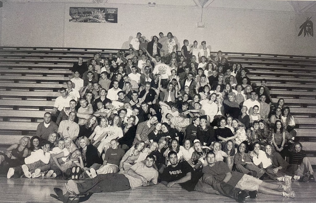 Large group of high school students sitting and standing in bleachers, smiling and posing together for a playful class photo in a gym.