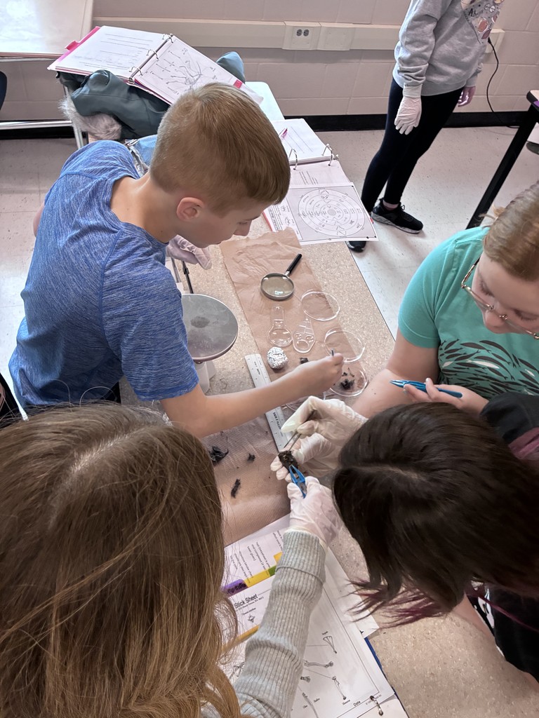 Students gather around a lab table, working together with magnifying tools, worksheets, and lab equipment in a science classroom.