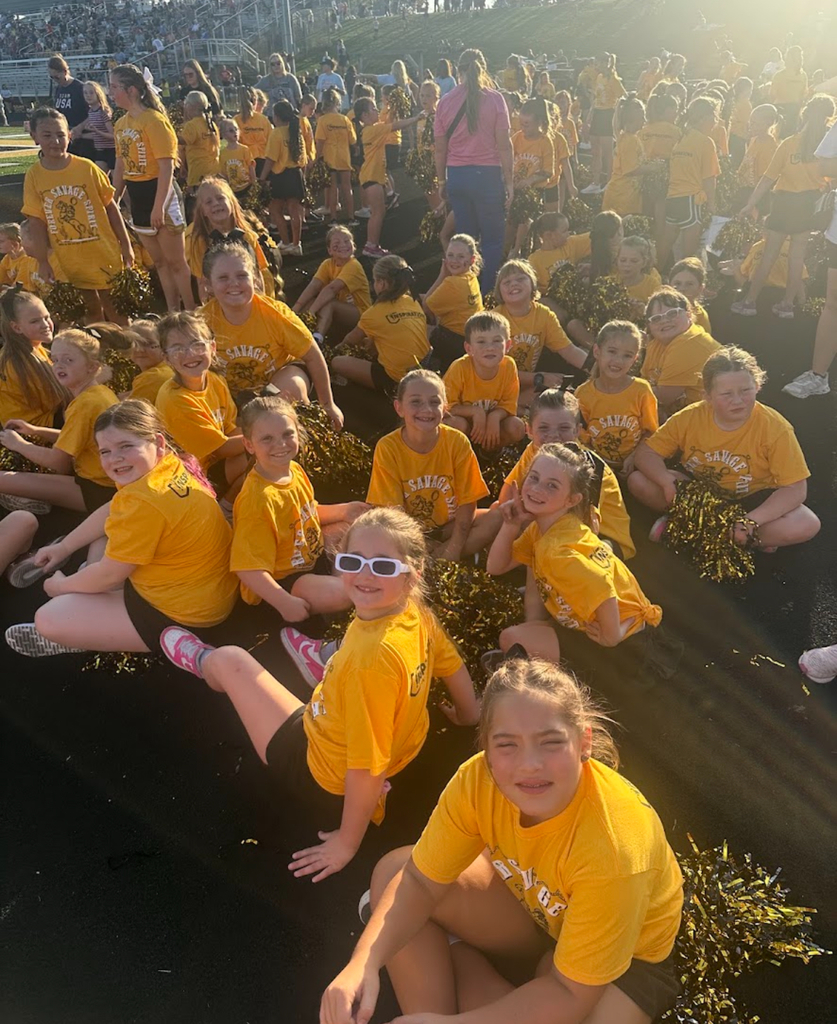 Group of elementary students wearing gold Savage Spirit Clinic shirts sitting on a track with pom-poms during a school spirit event.