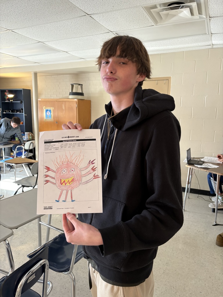 High school student holding a colored drawing of a monster created during a biology genetics activity, standing in a classroom.