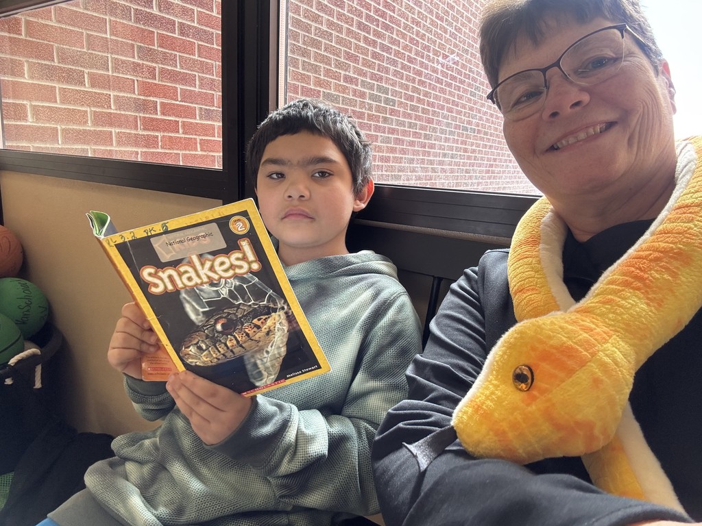 A student sitting next to a teacher indoors, holding and reading a book about snakes while the teacher smiles beside them.