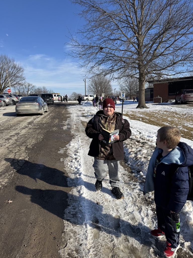 A student standing outside in the snow during parent pickup, holding a book and reading while waiting to go home.
