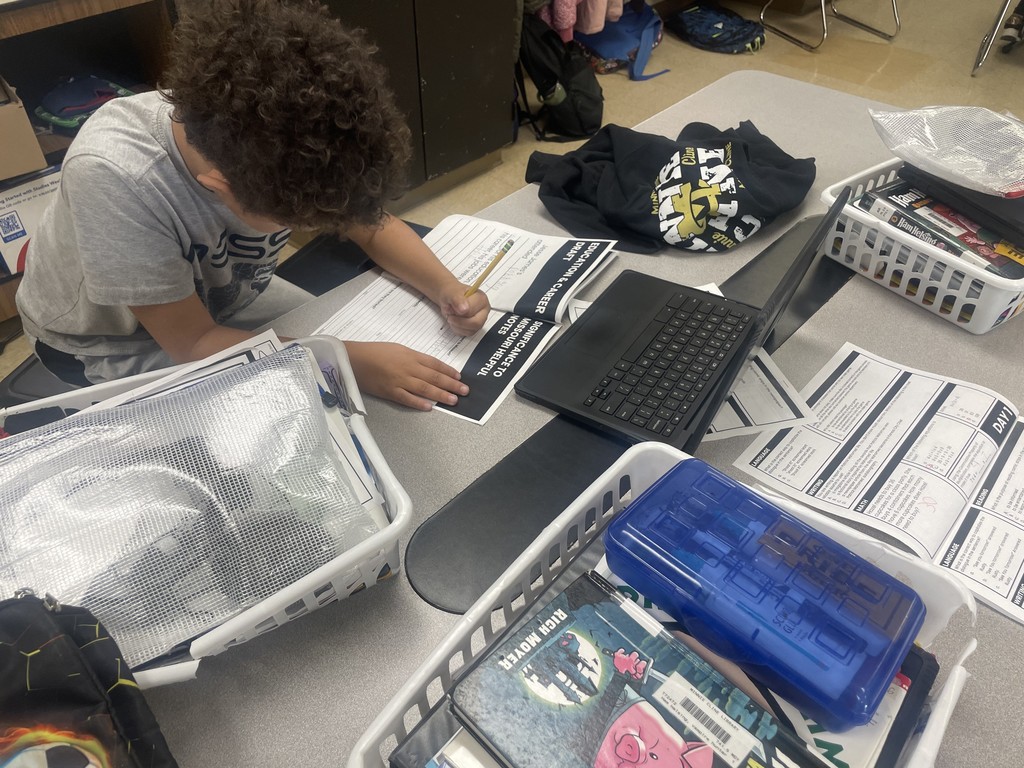 A student writing on a research worksheet while using a laptop at a classroom desk.