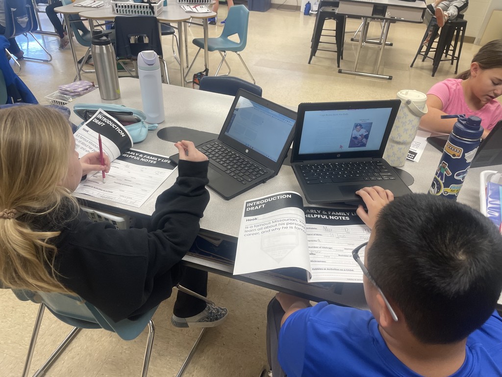 Several students working independently at tables with laptops, notebooks, and research materials.