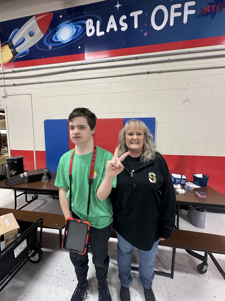 A high school student wearing a green shirt stands beside an adult in a school hallway, smiling for a photo.