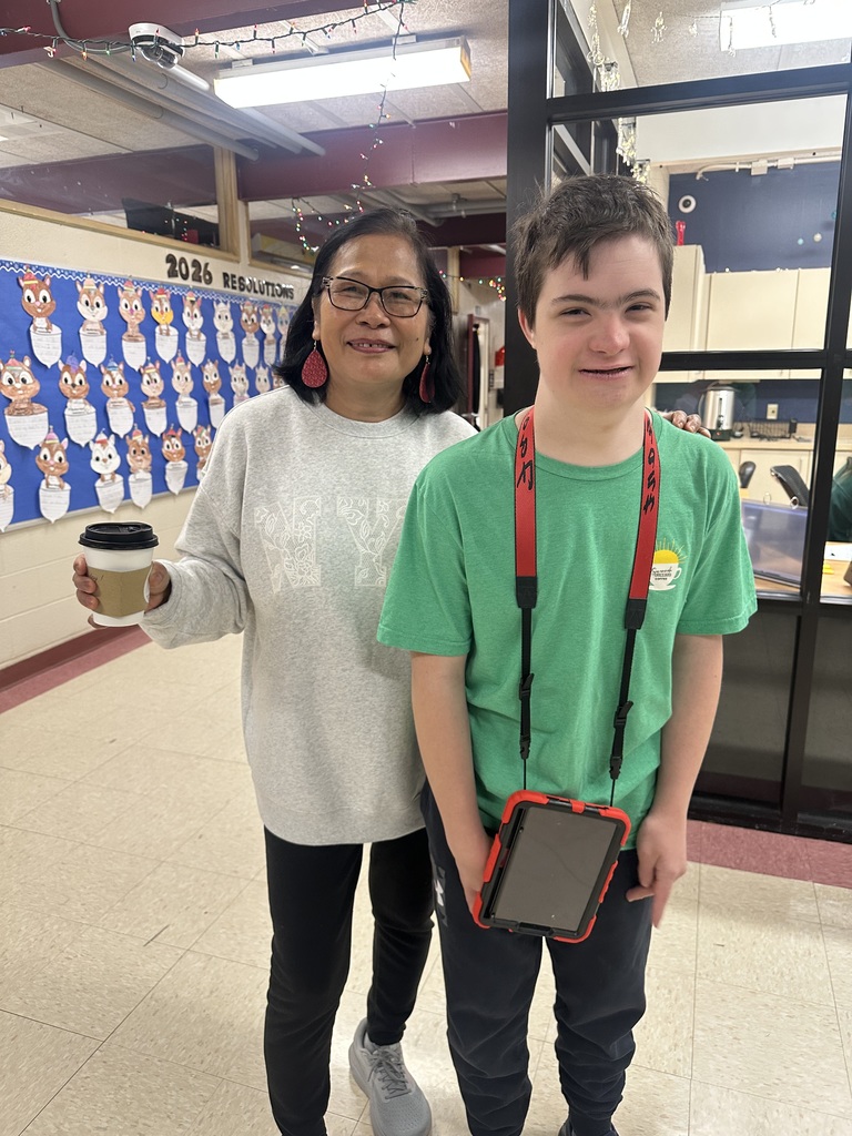 A high school student and staff member pose together inside a school cafeteria with a colorful wall mural behind them.