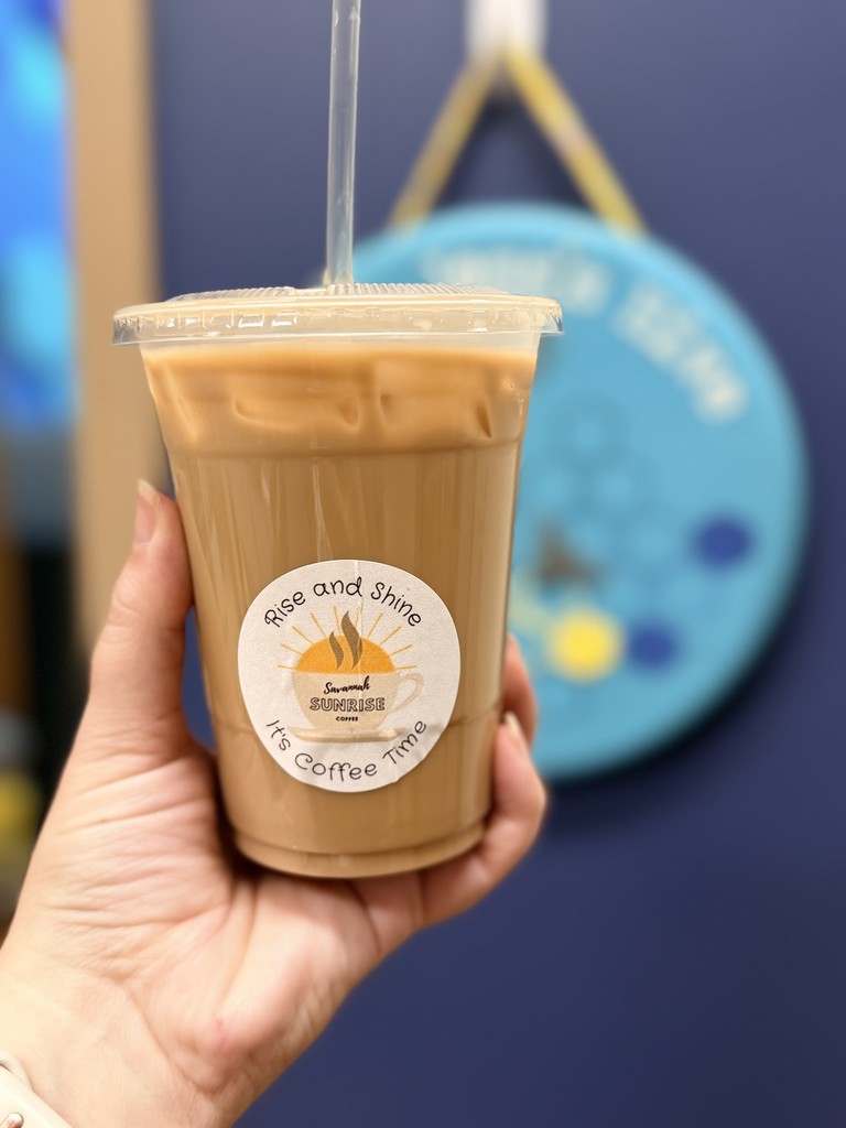 A hand holding an iced coffee cup with a “Rise and Shine, It’s Coffee Time” sticker.
