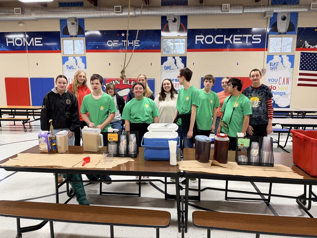 High school students wearing green shirts stand together behind a coffee service table labeled with different drink options.