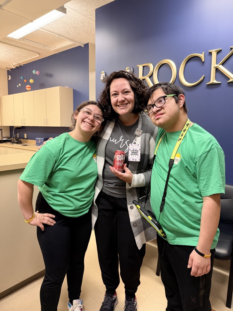 Two high school students in green shirts and a staff member pose together with a colorful wall mural behind them.