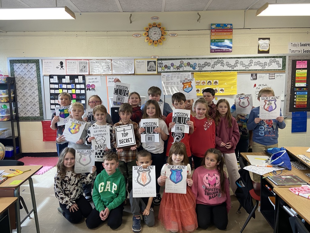 A full second-grade class poses in their classroom holding detective and secret-agent project pages created for a 100th day of school activity.