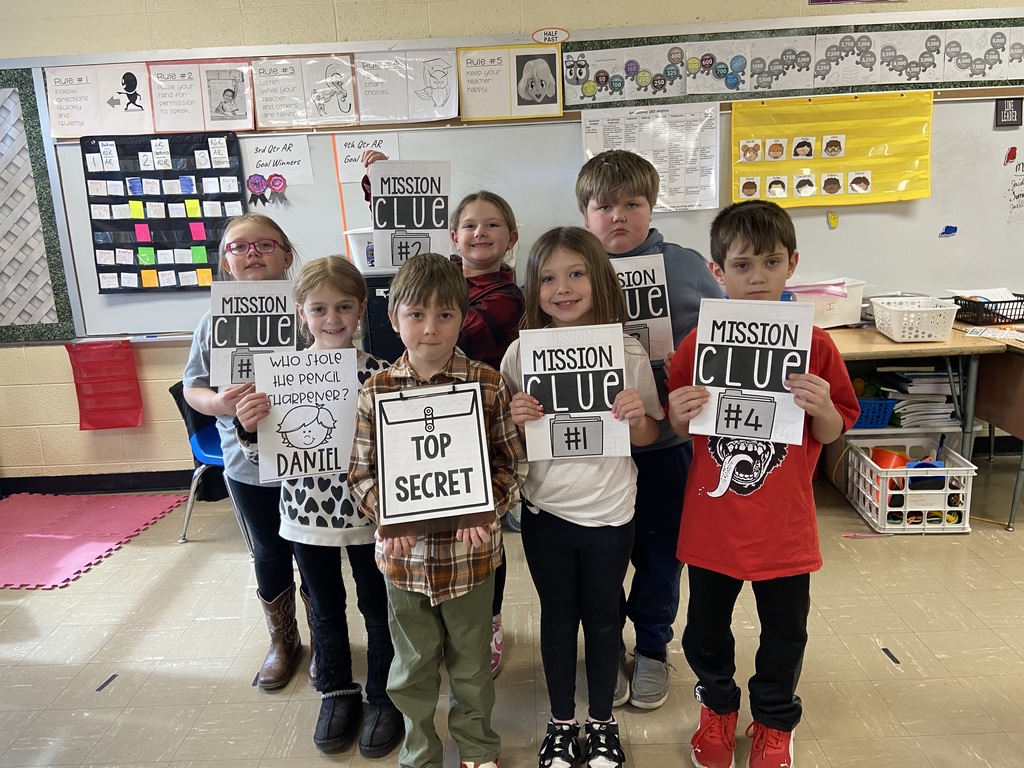 A small group of second-grade students stand in a classroom holding detective-themed signs that read “Mission Clue,” “Top Secret,” and “Secret Agent.”