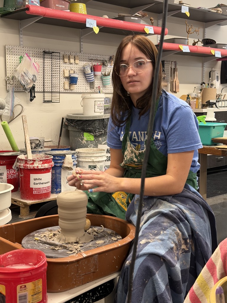 Student carefully forms a tall clay cylinder on a pottery wheel surrounded by tools and buckets.