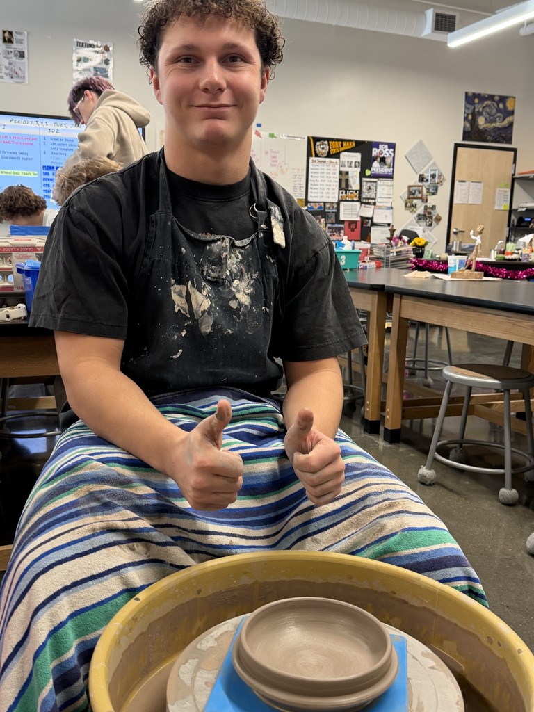 Student gives two thumbs up while seated at a pottery wheel with a freshly shaped clay bowl.