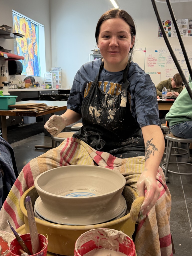 Student shapes a wide clay bowl on a spinning pottery wheel in art class.