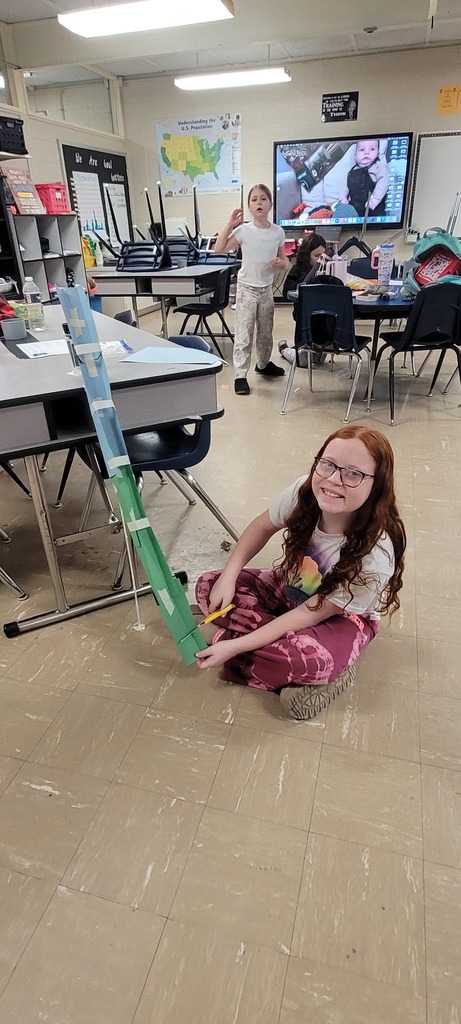 Student smiles while holding a green paper roller coaster model in a classroom.