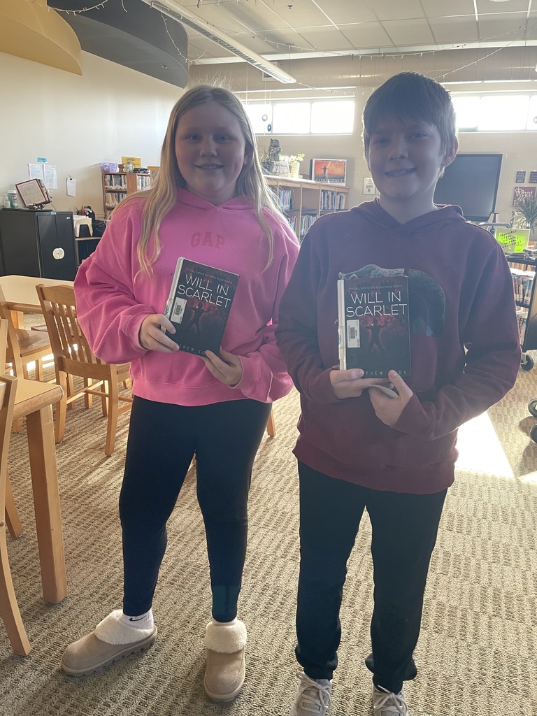 Two middle school students stand in the library holding copies of Will in Scarlet.