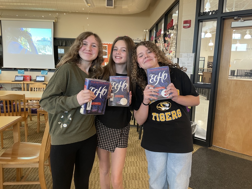 Three middle school girls stand in the library holding copies of Echo and smiling at the camera.