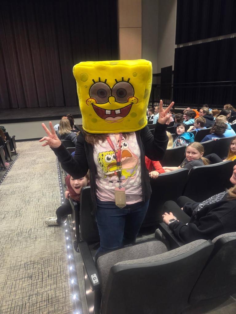 Elementary teacher wearing a SpongeBob costume stands in the SHS Performing Arts Center aisle while students sit in auditorium seats during a school field trip performance.