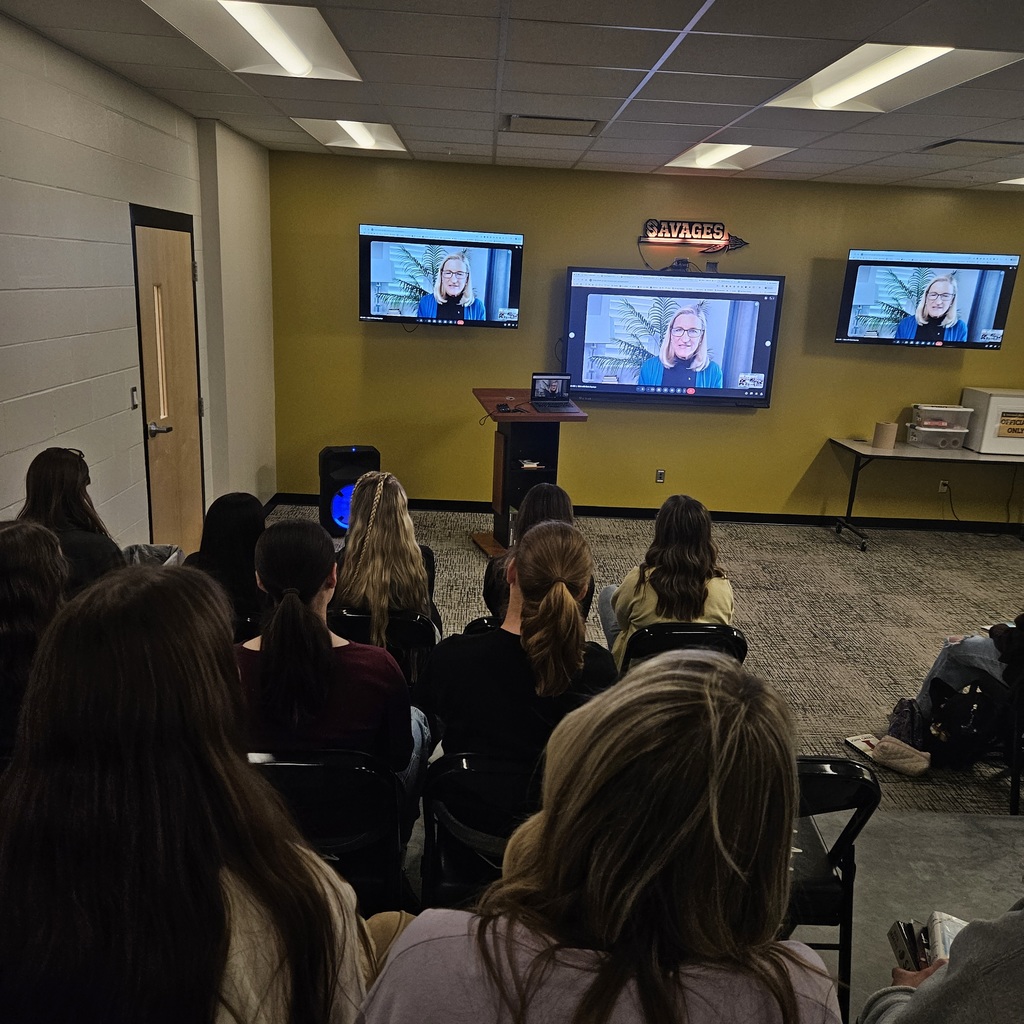Students sit in rows watching a virtual author speak on large screens at the front of a classroom.