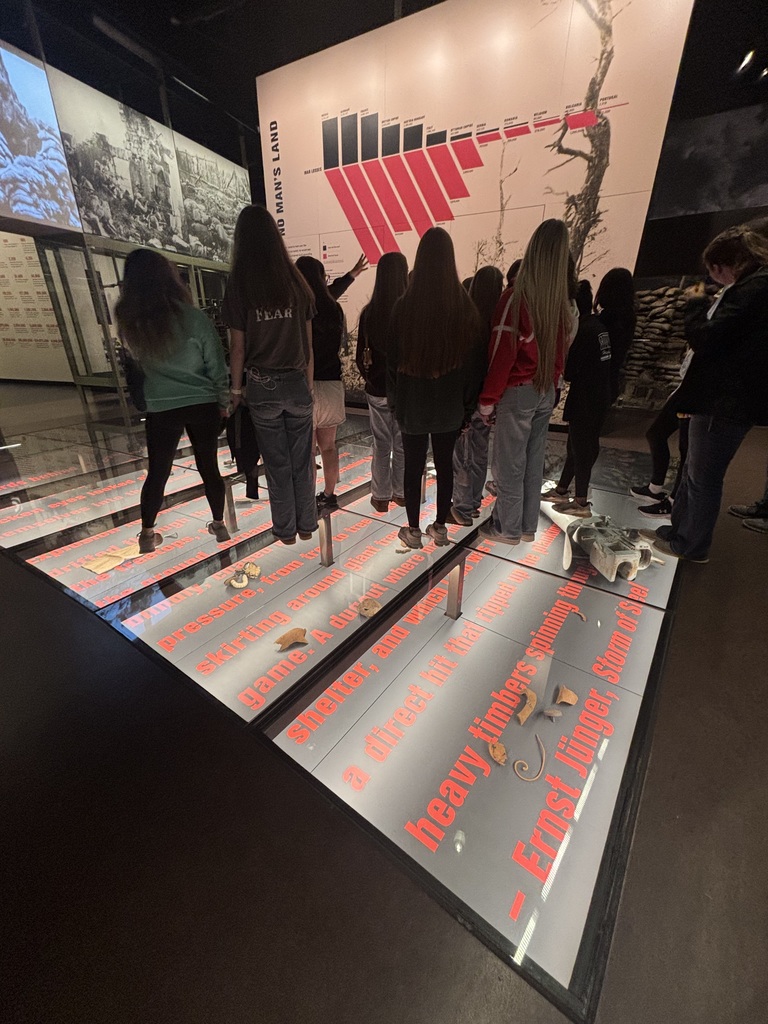Students listen to a museum guide beside a large “No Man’s Land” display while standing on a floor exhibit with projected text and artifacts.