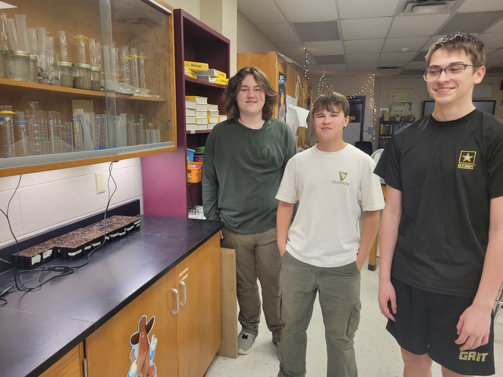 Three students stand beside trays of newly planted seeds under a grow light in the science classroom.
