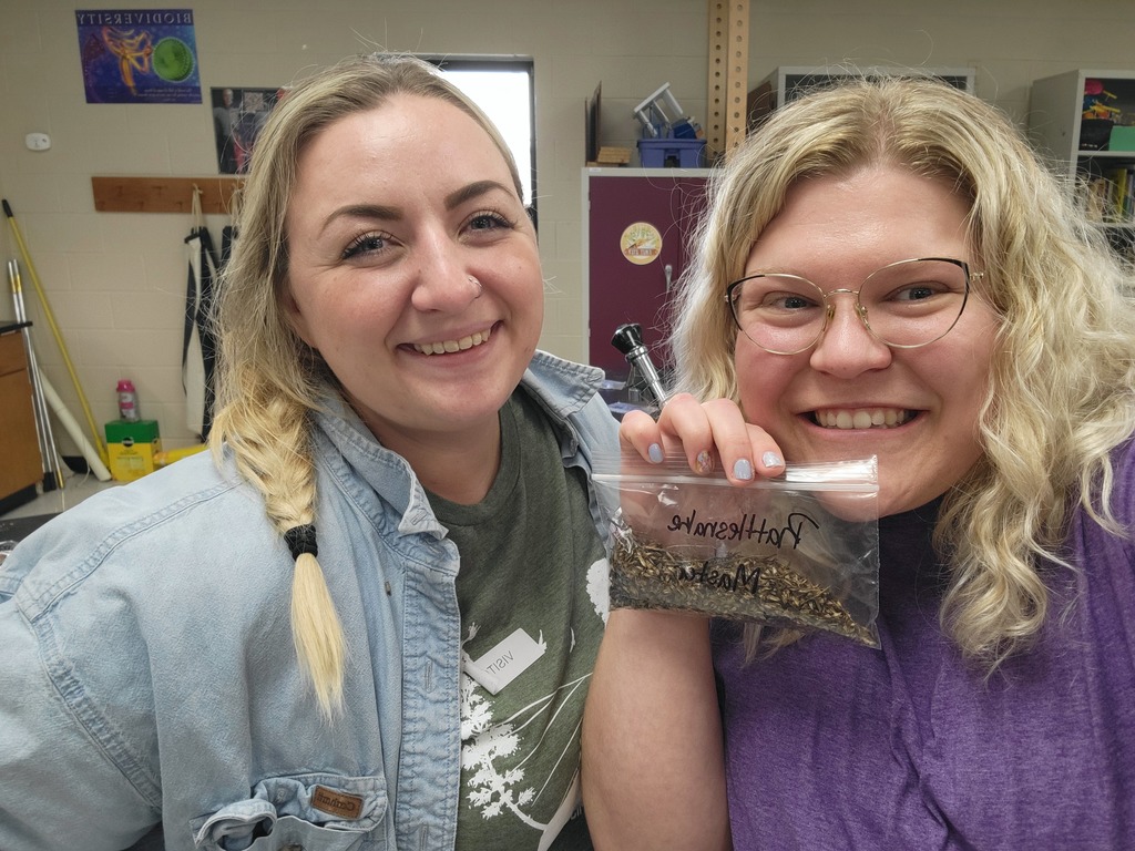 Two smiling staff members hold a labeled bag of prairie seeds collected for a classroom planting project.