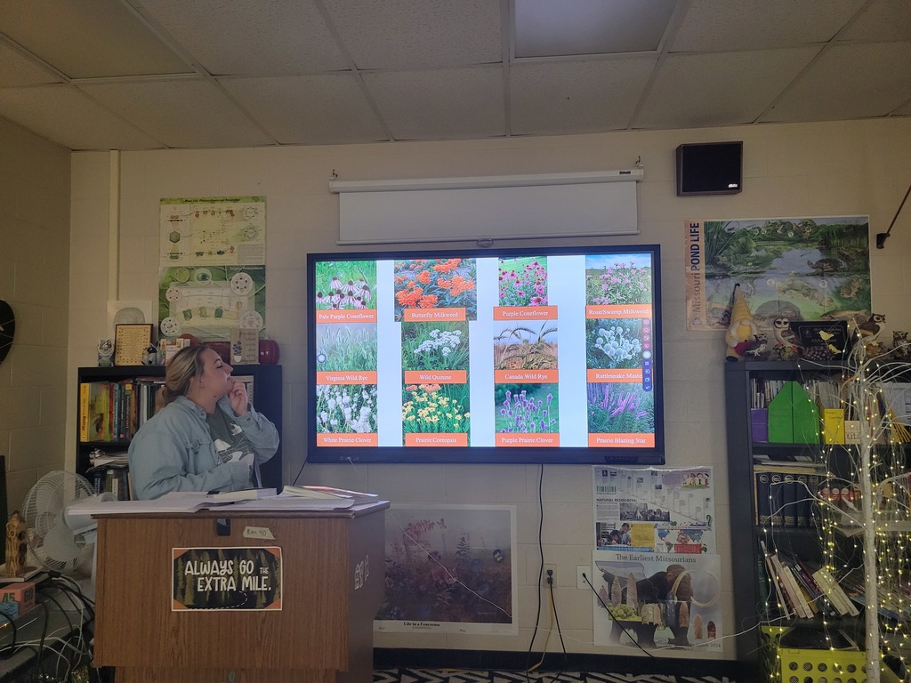 A guest presenter stands beside a classroom screen showing photos and names of Missouri native prairie plants.