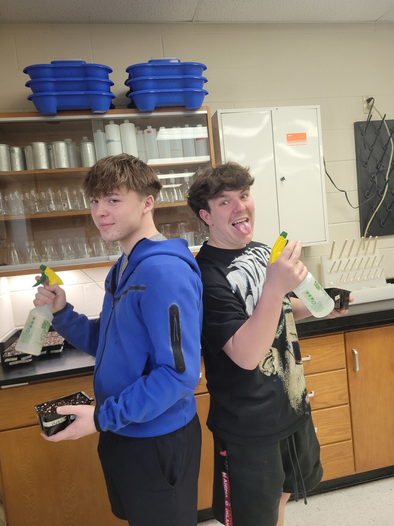 Two students stand back-to-back in a science lab holding spray bottles and seed trays as they help care for planted seeds.
