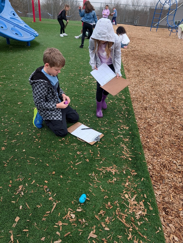 Two students pause over clipboards on the pavement while searching for and solving math problems from plastic eggs.