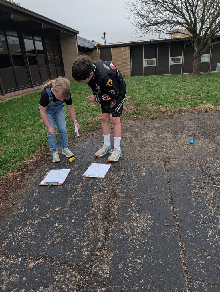 Two students work on math problems beside clipboards on the playground during an outdoor egg hunt activity.