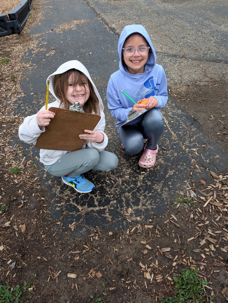 Two smiling students crouch on the blacktop holding clipboards, pencils, and plastic eggs during a math activity.