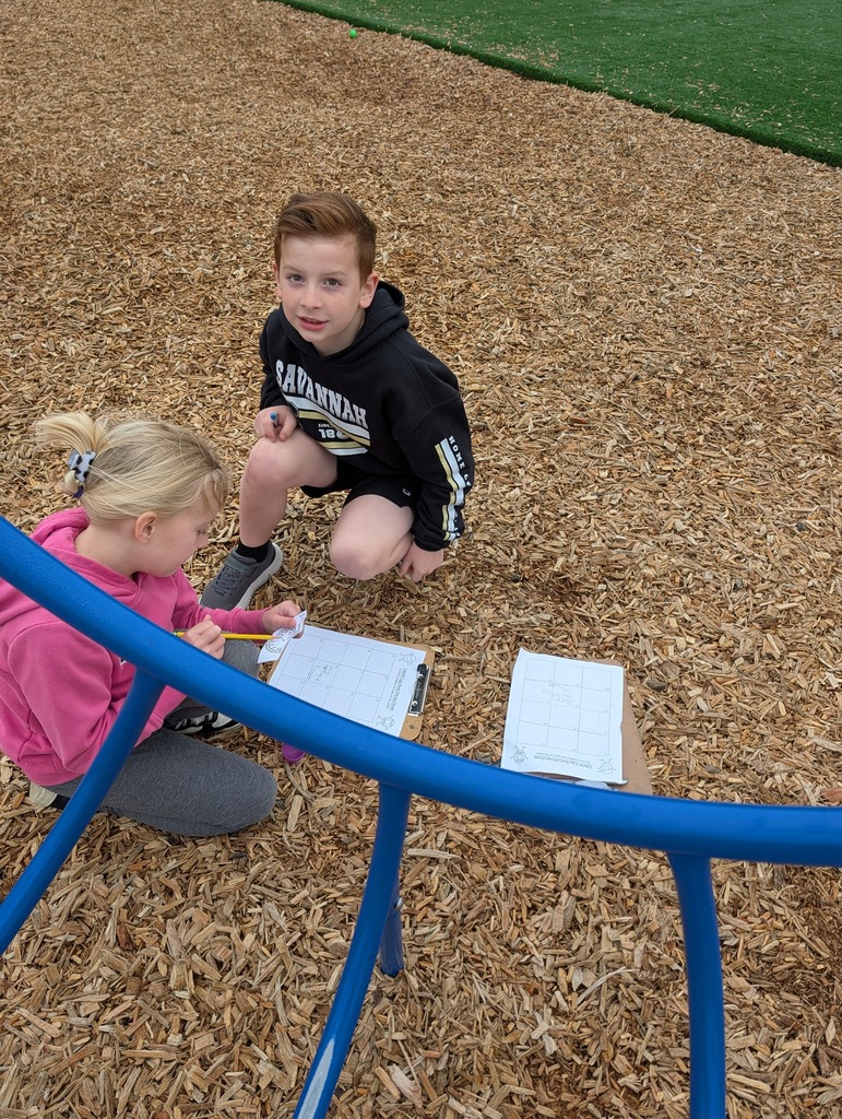 Two students kneel under playground equipment, solving a problem from a plastic egg with a clipboard nearby.