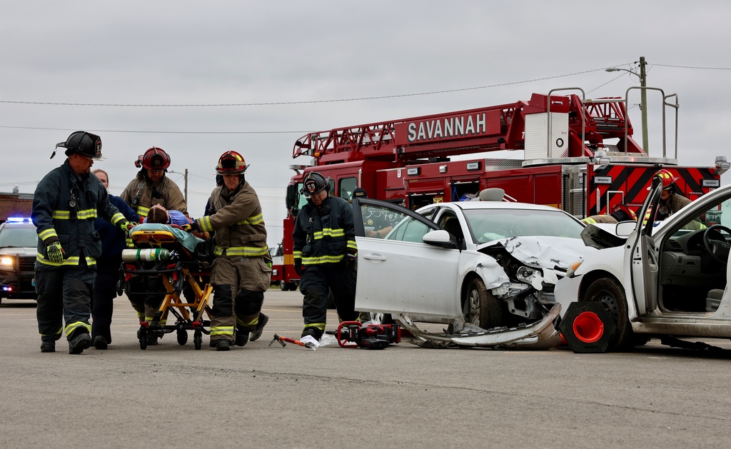 Firefighters transport a simulated crash victim on a stretcher near a damaged vehicle during the Docudrama.