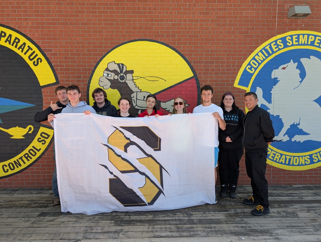 8 students stand in front of a brick wall holding a large white flag with the Savannah S with claw marks.  JROTC instructor Sgt. Addwel  stands to the side of the group. 